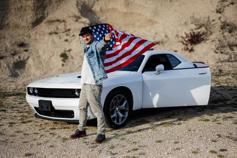 handsome man jeans jacket cap with usa flag near his white american muscle car career